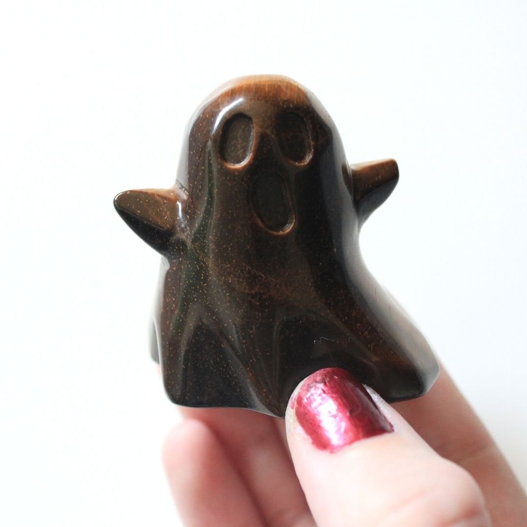 A photo of a small tiger's eye ghost figurine held in a hand against a white background. The ghost is carved from tiger's eye, a golden-brown gemstone with a characteristic chatoyancy, or shifting light effect. The ghost has simple facial features, with small, round eyes and a open mouth.