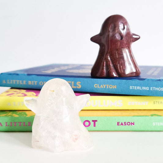 A photo of two small ghosts made of clear quartz and red ocean jasper, sitting on a stack of books. One ghost is made of clear quartz, a transparent mineral with a slight white tint, while the other ghost is made of red ocean jasper, a red and brown banded mineral. Both ghosts have simple facial features, with small, round eyes and a open mouth.