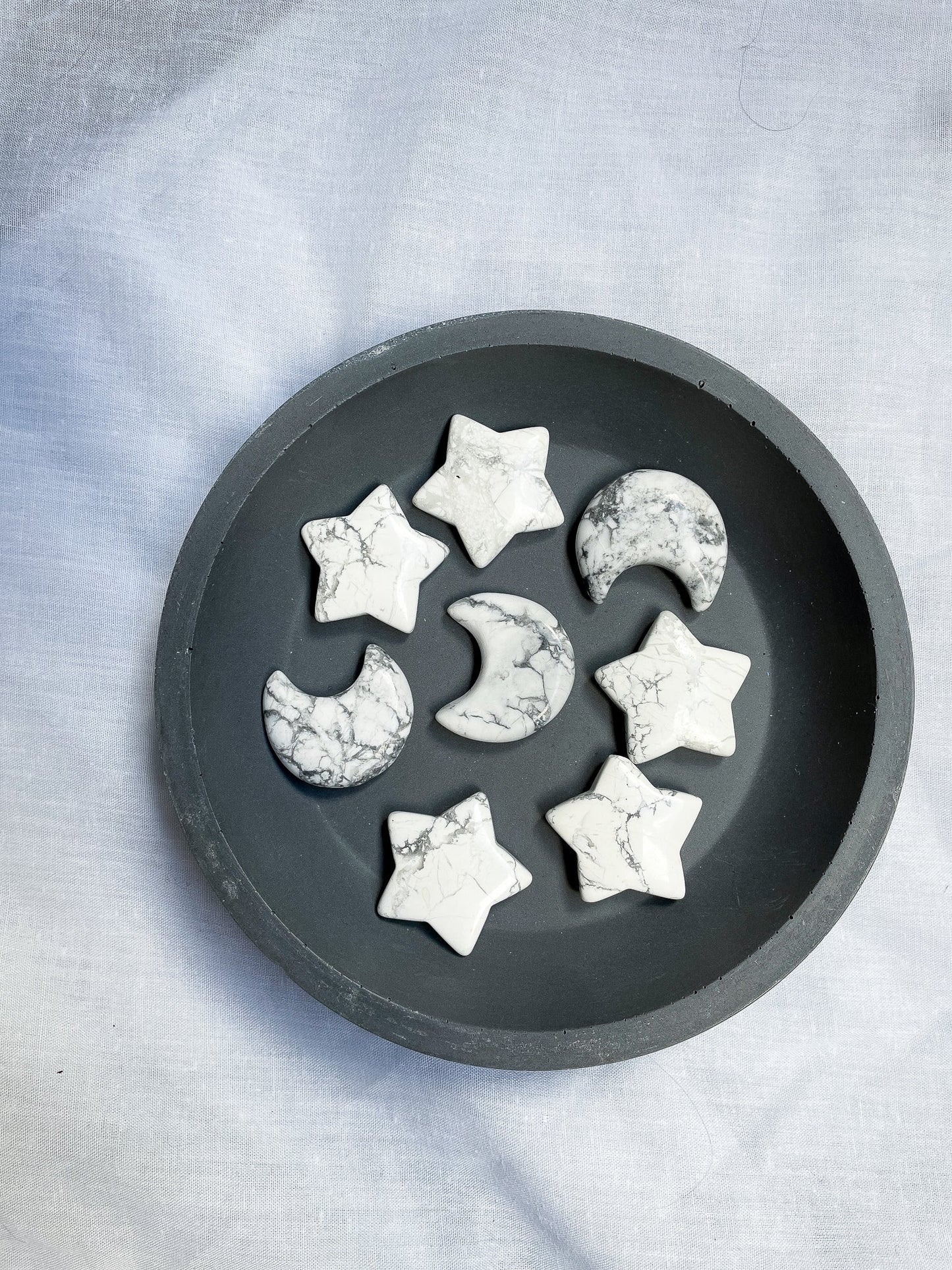A photo of howlite moon and star carvings resting in a dark grey dish, with a light grey background. The moon and star carvings are made from howlite, a mineral that is typically white or grey with grey, black, or brown veins or markings.