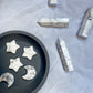 A photo of howlite moon and star carvings arranged in a grey bowl, along with several howlite points beside the bowl, against a light grey background.